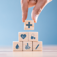 A hand laying cubes on which health-related symbols are drawn: a vertical cross, a heart, a pill, a drop of blood, a person in rehabilitation and a syringe.